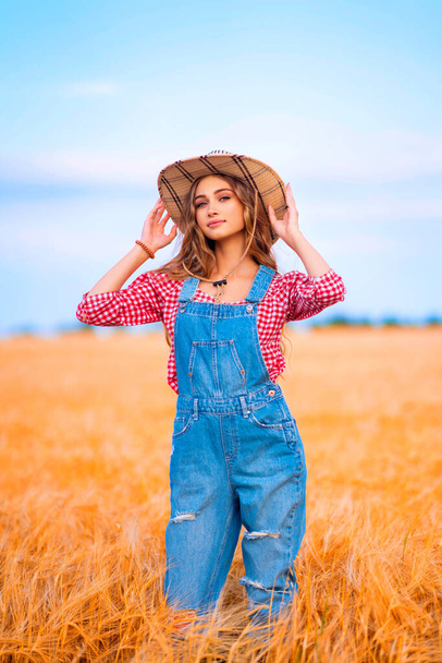若いです可愛いですブロンド女の子posingで分野の小麦.美しいですスタイリッシュなカウガールでわら帽子とデニムオーバーオールオン暖かいカラフルな田舎の写真. - 写真・画像