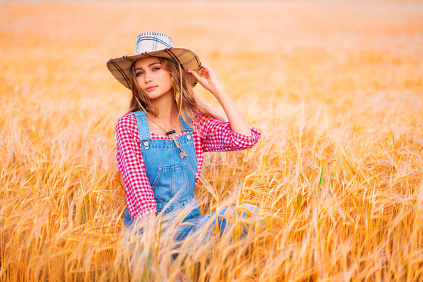 Joven chica rubia bonita posando en el campo de wheat.Beautiful vaquera con estilo en sombrero de paja y overoles de mezclilla en cálidas imágenes coloridas del campo. - Foto, imagen
