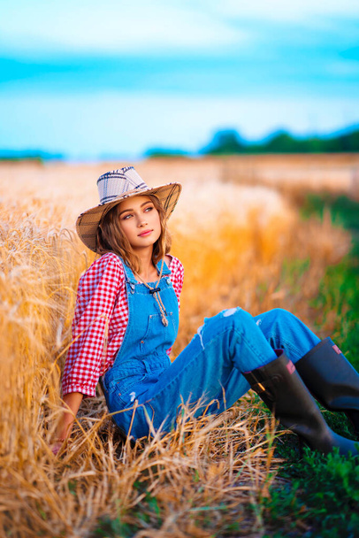 Genç, güzel sarışın kız buğday tarlasında poz veriyor. Hasır şapkalı ve kot pantolonlu güzel kovboy kız sıcak renkli kırsal resimlerde.. - Fotoğraf, Görsel