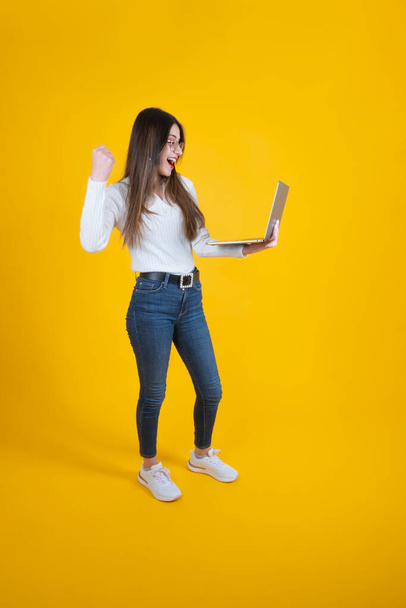Celebrating success,  full body length cheerful  woman celebrating success.  Caucasian  lady raising clenched fist, looking and holding modern laptop. Yellow studio background. Cheering victory. - Photo, image