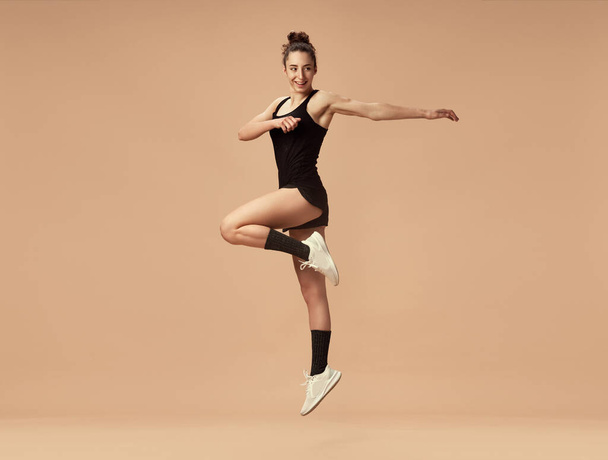 Dynamic image of sportive young girl in black sportswear training, jumping against light brown studio background. Building muscles. Concept of sportive lifestyle, beauty, body care, fitness, health - Foto, afbeelding