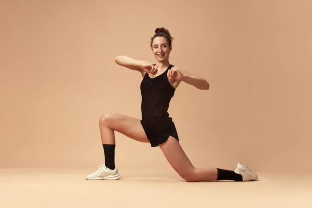 Flexible, sportive, smiling young girl in sportswear training, stretching against light brown studio background. Workout. Concept of sportive lifestyle, beauty, body care, fitness, health - Фото, зображення