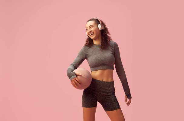Young smiling girl with slim body in gray sportswear posing in headphones and basketball ball against pink studio background. Concept of sportive lifestyle, beauty, body care, fitness, health - Foto, afbeelding