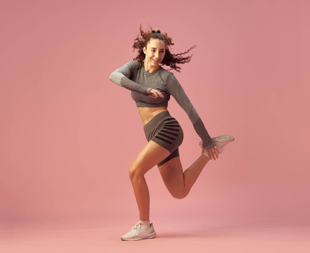 Gri spor giyimli genç kızın dinamik görüntüsü. Pembe stüdyo geçmişine zıplıyor. Zayıf, formda bir vücut. Spor yaşam tarzı, güzellik, vücut bakımı, fitness, sağlık - Fotoğraf, Görsel