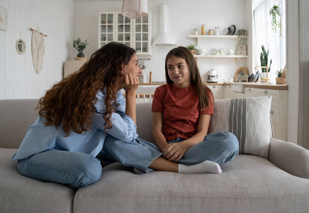 Happy good-natured mother communicates with teenage girl listening to dreams about future or stories about school life. Smiling optimistic european woman sits with daughter on sofa in home interior - Foto, Bild