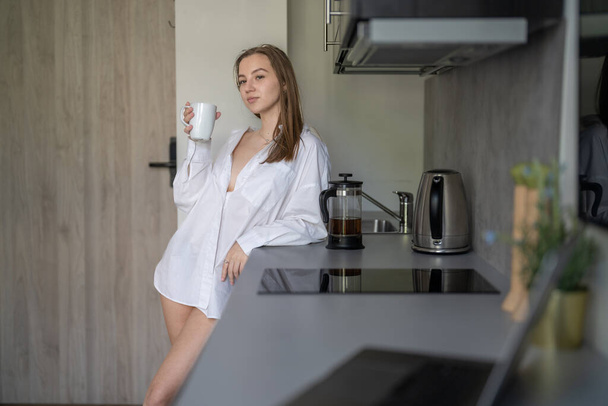 美しい若い女の子がキッチンに立ってコーヒーを飲んでいます。朝のルーチン。白いシャツを着て。机の上のブリュリーノートパソコンと携帯電話. - 写真・画像