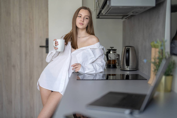 Beautiful Young Girl está de pie en la cocina y tomando café. Buenos días Rutina. Con camisas blancas. Ordenador portátil borroso y teléfono móvil en el escritorio. - Foto, Imagen