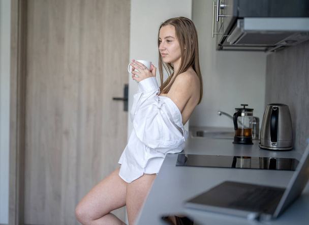 Beautiful Young Girl está de pie en la cocina y tomando café. Buenos días Rutina. Con camisas blancas. Ordenador portátil borroso y teléfono móvil en el escritorio. - Foto, imagen