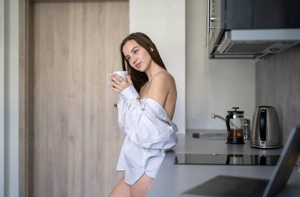 Kaunis nuori tyttö seisoo keittiössä ja juo kahvia. Huomenta, Rutiini. Valkoisissa paidoissa. Sumea kannettava tietokone ja matkapuhelin pöydällä. - Valokuva, kuva