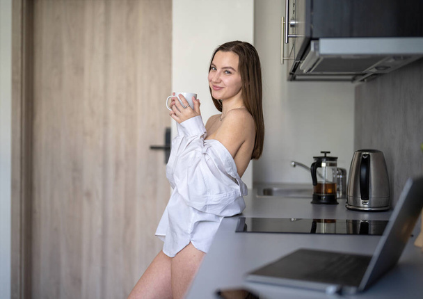 Menina bonita está de pé na cozinha e beber café. Rotina da manhã. Vestindo camisas brancas. Laptop embaçado e telefone celular na mesa. - Foto, Imagem