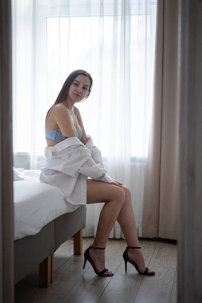 Beautiful Young Girl Wearing Blue Bra and White Shirt is Sitting on the Bed in the Bedroom. Interior - Foto, Imagen