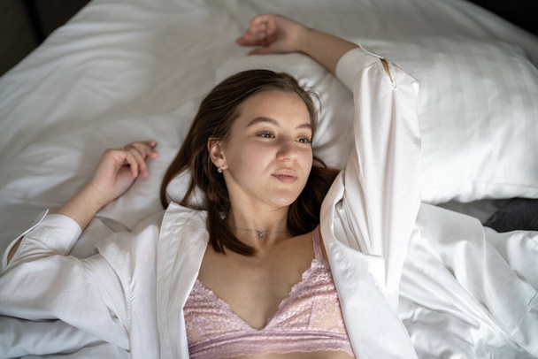 Όμορφη νεαρή κοπέλα είναι ξαπλωμένη στο κρεβάτι στην κρεβατοκάμαρα. Φορώντας λευκά πουκάμισα. Πορτρέτο - Φωτογραφία, εικόνα