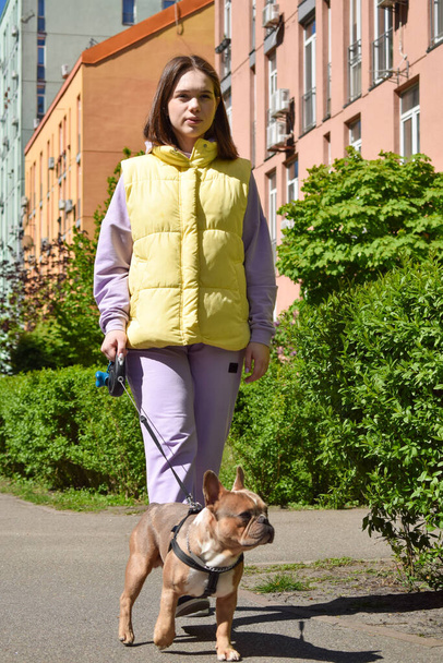 Hermosa mujer joven paseando encantador perro pequeño en el área urbana en el día soleado. Obediente perro animal doméstico paseando con el dueño de la mascota al aire libre. Distrito residencial en segundo plano. Ciudad concepto de estilo de vida - Foto, Imagen