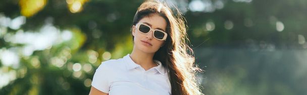 charming and young woman with brunette long hair standing in white polo shirt and trendy sunglasses near blurred and green palm trees in Miami, sunny day, travel, vacation, banner, sun-kissed  - Photo, Image