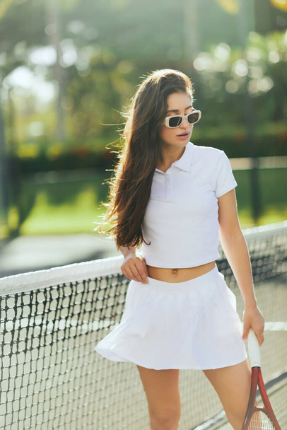 tennis court in Miami, sporty young woman with brunette long hair standing in white outfit and sunglasses while holding racket near tennis net, blurred background, iconic city, Florida  - Φωτογραφία, εικόνα