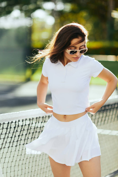 portrait of pretty young woman with brunette long hair standing in white outfit and sunglasses near tennis net, blurred background, wind, tennis court in Miami, iconic city, female player, Florida  - Photo, Image