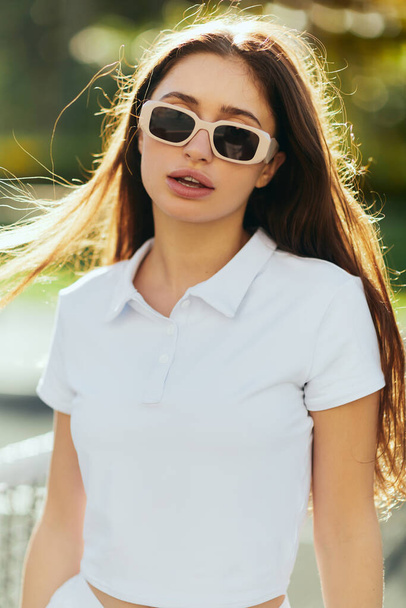 portré bájos fiatal nő barna hosszú haj áll fehér póló és napszemüveg közelében tenisz háló, homályos háttér, szél, teniszpálya Miami, ikonikus város, Florida, napsütéses nap  - Fotó, kép
