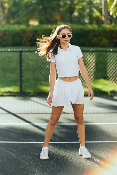 urheilullinen nuori nainen ruskeaverikkö hiukset poninhäntä ja aurinkolasit seisoo valkoinen asu tennishame lämmittäessään tenniskenttä hämärtynyt tausta, Miami, Florida - Valokuva, kuva