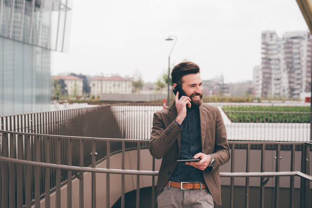 Joven contemporáneo barbudo moderno hombre de negocios al aire libre hablando smartphone celebración tableta - Foto, Imagen