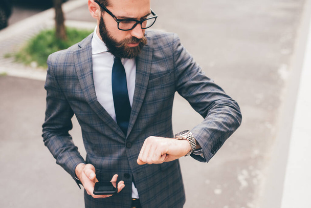 Молодий професійний елегантний бородатий бізнесмен на відкритому повітрі перевіряє час на наручних годинниках
 - Фото, зображення