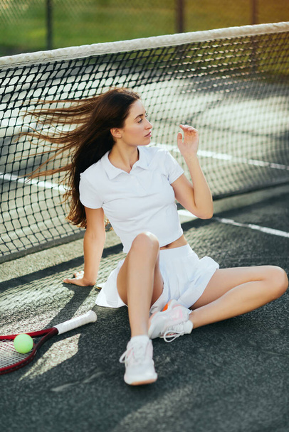 female tennis player sitting against wind, young woman with long hair sitting in white outfit near racket with ball and tennis net, blurred background, Miami, iconic city, tennis court, downtime - Photo, Image