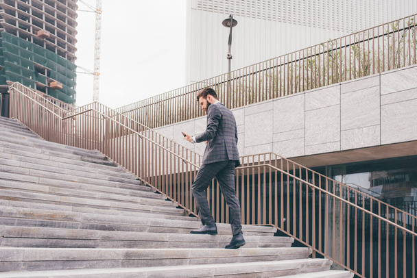 Jovem barbudo elegante homem de negócios indo para cima andando ao ar livre usando smartphone - Foto, Imagem