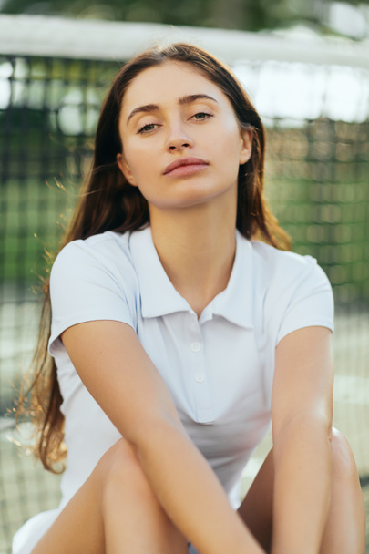 portrait of female tennis player with brunette long hair wearing white polo shirt and looking at camera after training on tennis court, tennis net on blurred background, Miami, Florida - Foto, Bild