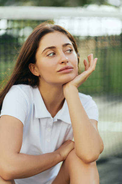 tennis court in Miami, portrait of dreamy female tennis player with brunette hair wearing white polo shirt and looking away after training, tennis net on blurred background, Florida - Photo, Image