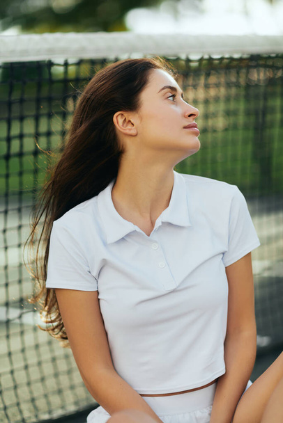 tennis court in Miami, portrait of distracted female tennis player with brunette hair wearing white polo shirt and looking away after training, tennis net on blurred background, Florida - Φωτογραφία, εικόνα