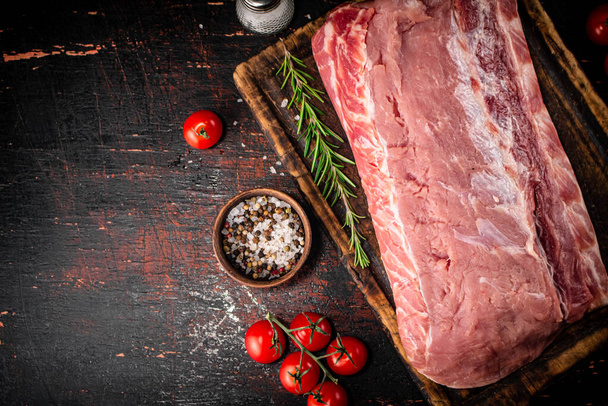 A piece of raw pork on a cutting board with tomatoes, spices and rosemary. On a rustic dark background. High quality photo - Photo, Image
