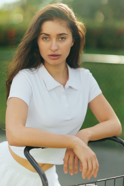 pretty tennis player, sporty young woman with brunette hair standing in white polo shirt near tennis cart, blurred green background, looking at camera, tennis court in Miami  - Foto, imagen