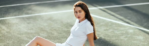 female tennis player resting after game, young woman sitting in white outfit on asphalt and looking at camera, blurred background, Miami, tennis court, downtime, polo shirt, banner  - Photo, Image