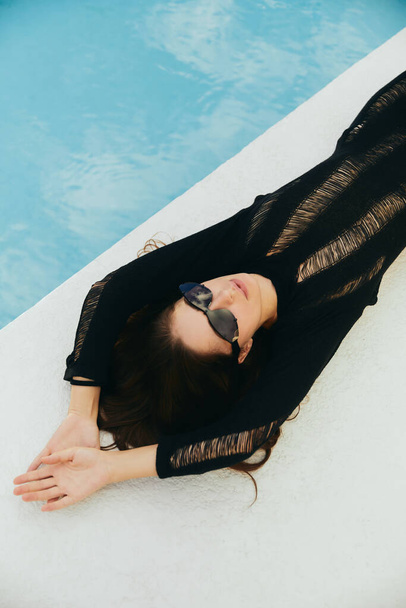 πειρασμός, πολυτελές θέρετρο, σέξι γυναίκα με μαυρισμένο δέρμα σε μαύρο πλεκτό φόρεμα και γυαλιά ηλίου δίπλα σε εξωτερική πισίνα με μπλε νερά στο Μαϊάμι, top view  - Φωτογραφία, εικόνα