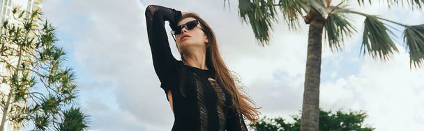 resort de lujo, mujer morena sexy con piel bronceada en vestido de punto negro y gafas de sol de pie contra las palmeras y el cielo azul en Miami, escapada de verano, bandera - Foto, Imagen