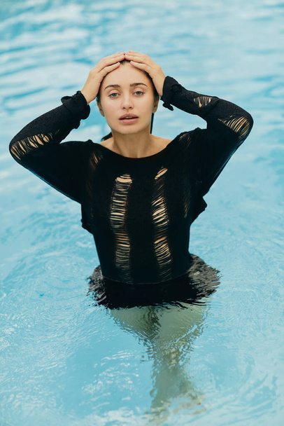 geen make-up look, aantrekkelijke en sexy brunette vrouw in zwarte gebreide outfit poseren in het buitenzwembad tijdens vakantie in Miami, verleidelijk, luxe resort, Florida  - Foto, afbeelding