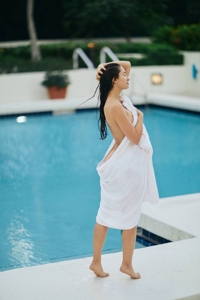 junge sexy Frau mit nassen Haaren, die in weißes Handtuch gehüllt neben einem Swimmingpool mit schimmerndem Wasser in Miami steht, Sommerurlaub, Jugend, Erholung am Pool, Urlaubsmodus  - Foto, Bild