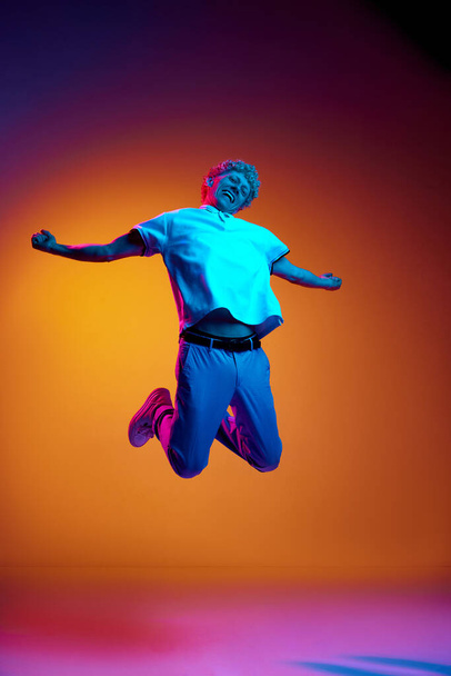 Full-length portrait of mature an in casual clothes jumping against orange studio background in blue neon light. Excited and happy. Concept of human emotions, lifestyle, youth, facial expression - Photo, Image