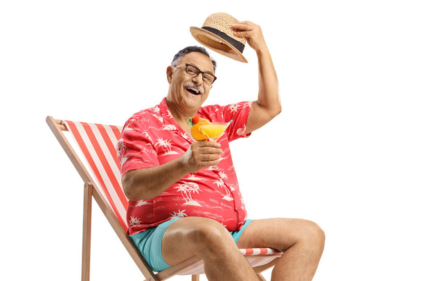 Mature male tourist toasting with a cocktail on a beach chair and greeting with his hat isolated on white background - Photo, image