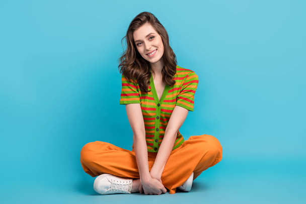Full size photo of good mood gorgeous woman dressed orange pants colorful cardigan sitting legs folded isolated on blue color background. - 写真・画像