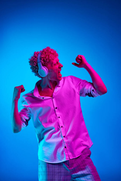Portrait of emotive mature man in white shirt listening to music in headphones against blue studio background in pink neon light. Concept of human emotions, lifestyle, youth, facial expression - Foto, Imagen