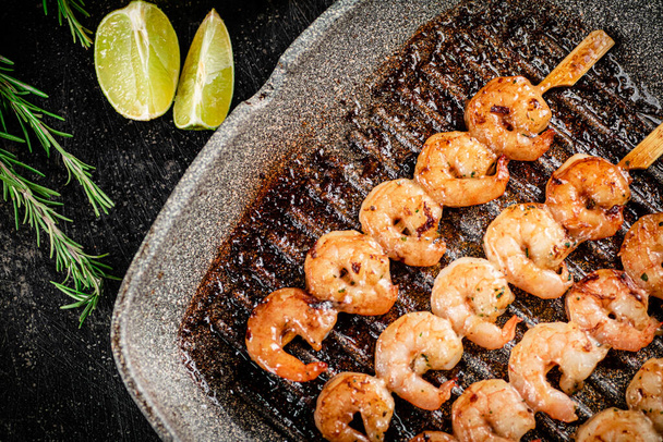 Grilled shrimp on skewers in a frying pan with rosemary and pieces of lime. On a black background. High quality photo - Photo, Image