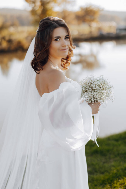 Portrait of a beautiful young bride in an elegant white dress with a long veil and a bouquet of gypsophila near the lake in soft light. The bride is looking at the camera. Portrait of the bride. - Foto, Bild