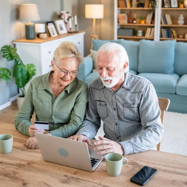 couple mature senior women and man husband and wife sit at home use credit or debit card for online shopping browse internet stores buying stuff use laptop computer real people copy space - Photo, image