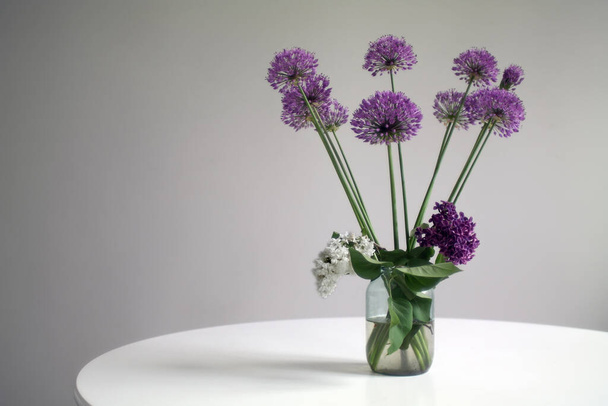 白い丸いテーブルの上にガラス瓶に巨大なタマネギの花とライラックの花束。選択的焦点. - 写真・画像