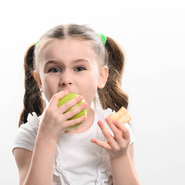 Küçük bir kız, beyaz arka planda bir elma ve patates kızartması tutuyor. Bir çocuğun hayatında sağlıklı ve sağlıksız yiyecek seçimi kavramı.. - Fotoğraf, Görsel