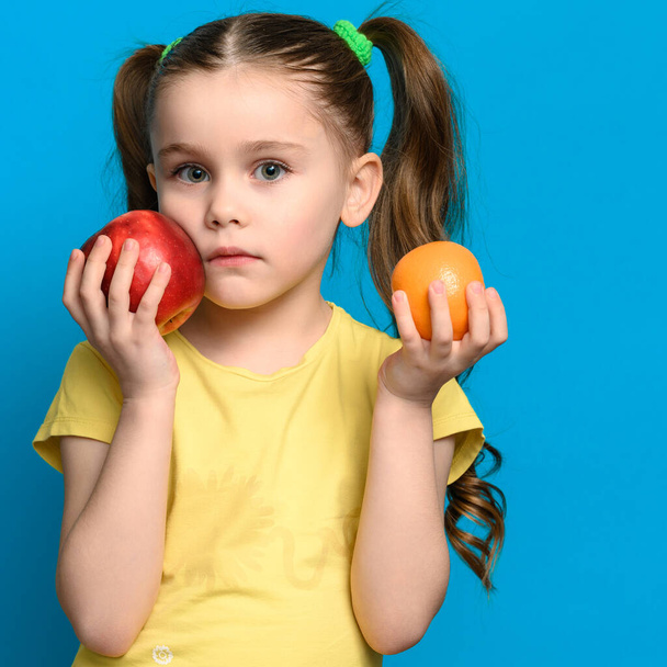 Auf blauem Hintergrund hält das Mädchen einen Apfel und eine Mandarine an der Wange, Früchte für Babynahrung. - Foto, Bild