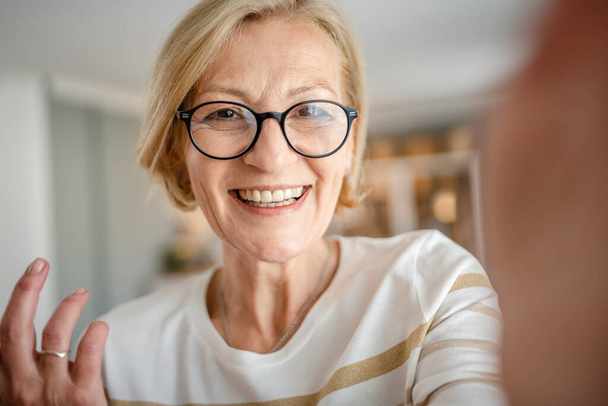 Portret van een volwassen blonde blanke vrouw met bril thuis vrolijke glimlach op zoek naar de camera zelfverzekerde dragen trui in lichte kamer user generated content zelfportret selfie ugc kopieerruimte - Foto, afbeelding