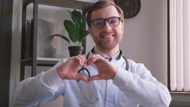 彼の現代的なオフィスに立っているヨーロッパの外観の若い肯定的な医師の肖像画は、カメラで微笑んだ。心臓内科医は自分の手で心臓のジェスチャーを示しています - 映像、動画