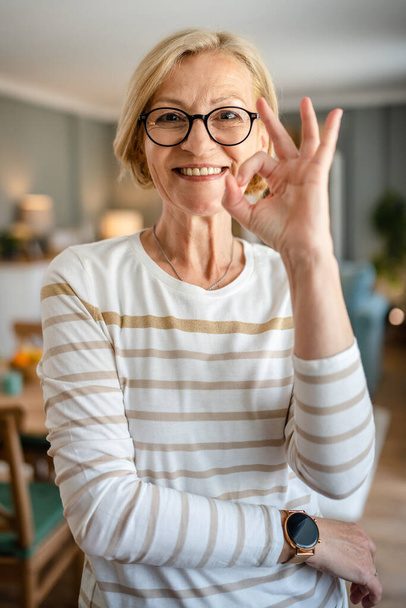 Πορτρέτο μιας ώριμης ξανθιάς καυκάσιας γυναίκας με γυαλιά στο σπίτι χαρούμενο χαμόγελο κοιτάζοντας προς την κάμερα αυτοπεποίθηση φορούν πουλόβερ σε φωτεινό χώρο αντίγραφο δωμάτιο - Φωτογραφία, εικόνα