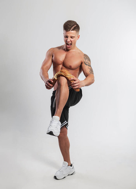 chladný emocionální silný atletický muž s svalnatým tělem v kraťasech a teniskách láme bagetový chléb o koleno na bílém pozadí. Zdravé jídlo a pečení, koncept - Fotografie, Obrázek